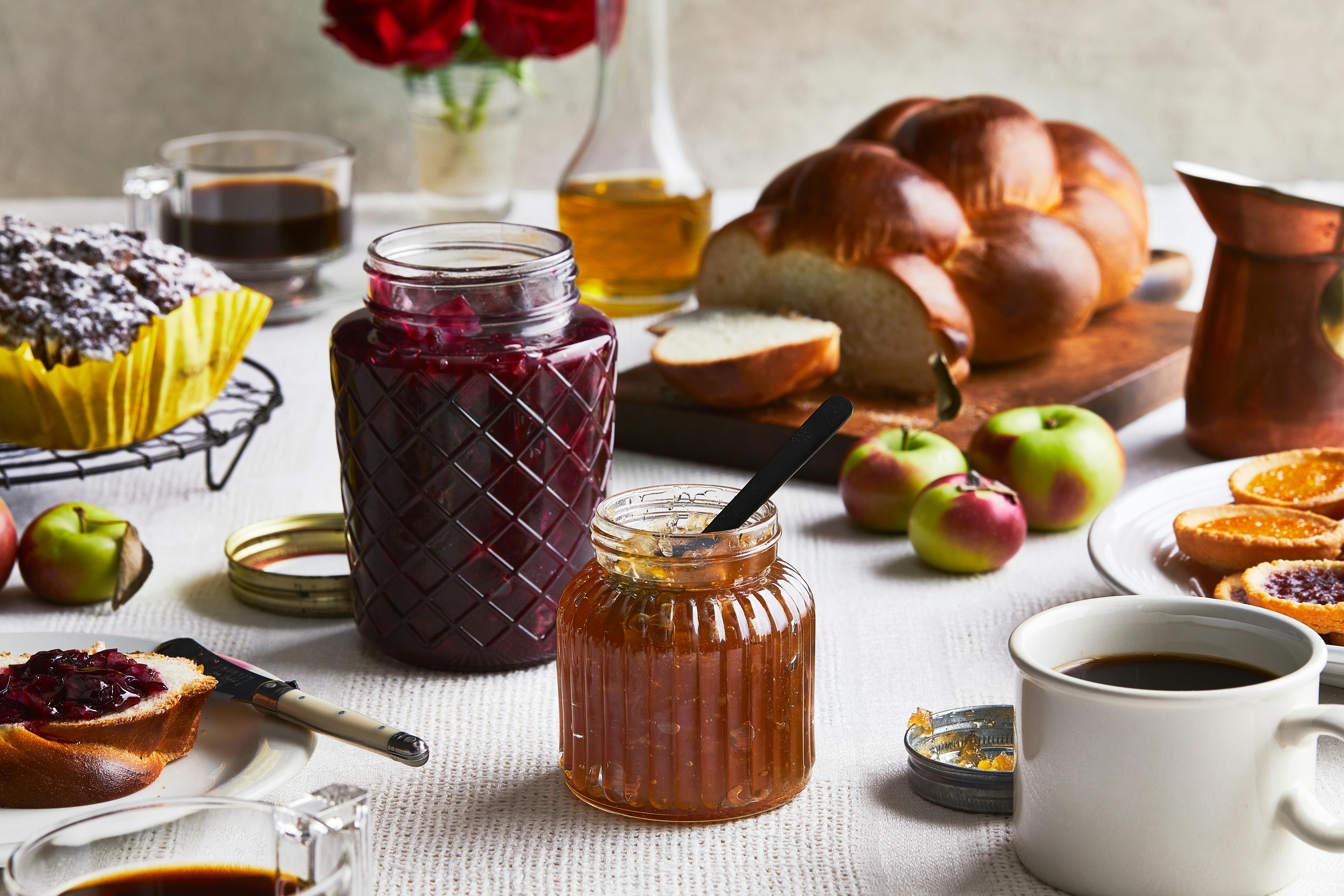 Rose and apple jams in glass jars on a table full of challah, apples, cake and cookies. 