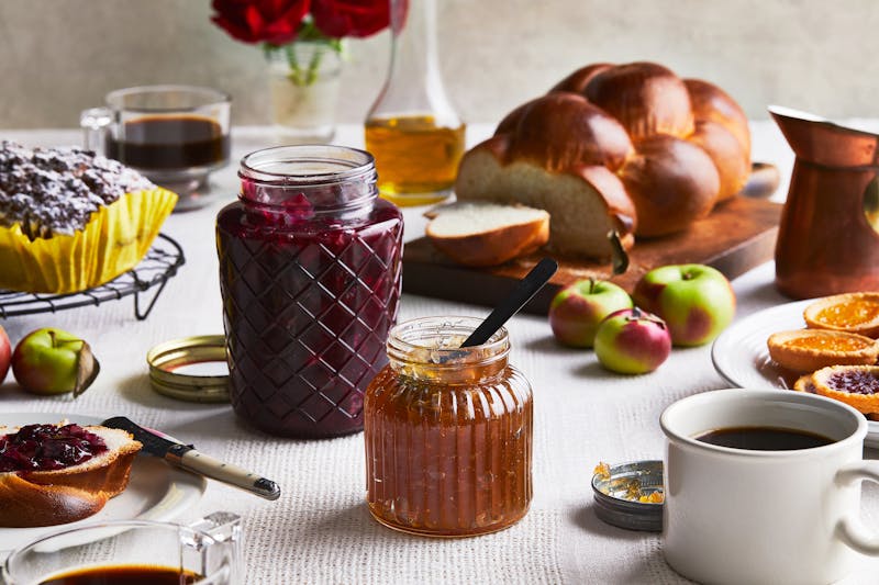 Rose and apple jams in glass jars on a table full of challah, apples, cake and cookies. 
