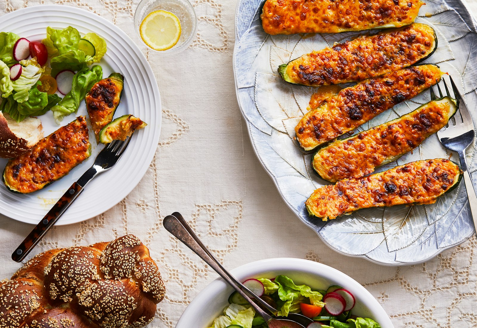A white tablecloth covered table is laden with an oval dish of stuffed zucchini in the upper right hand corner, and bowl of salad and loaf of challah in the upper and bottom left of the photo. 