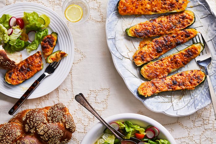 A white tablecloth covered table is laden with an oval dish of stuffed zucchini in the upper right hand corner, and bowl of salad and loaf of challah in the upper and bottom left of the photo.