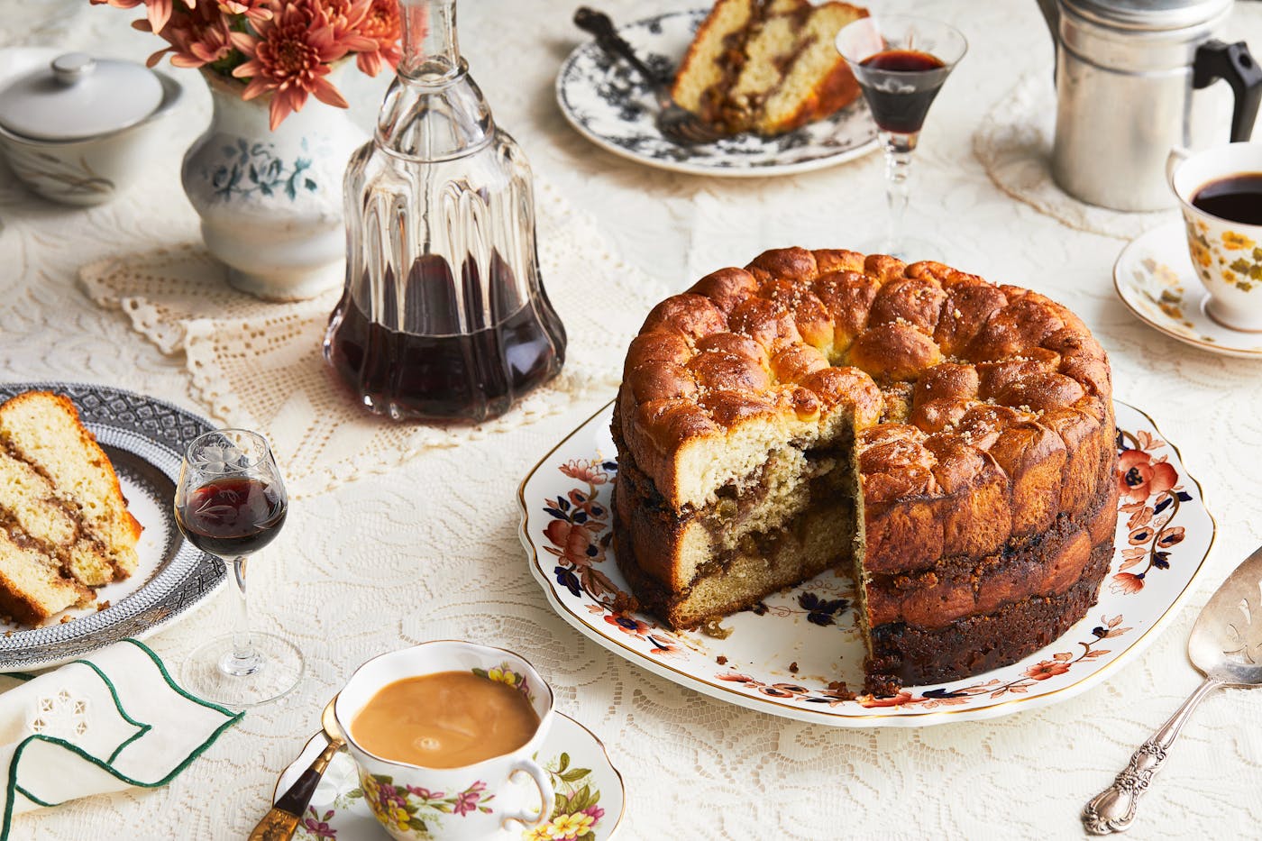An opened aranygaluska cake sits on a platter, surrounded by cups of coffee, a bottle of cherry liqueur and a plate with a slice of cake. 
