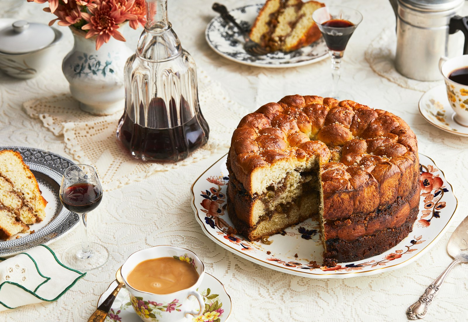 An opened aranygaluska cake sits on a platter, surrounded by cups of coffee, a bottle of cherry liqueur and a plate with a slice of cake. 
