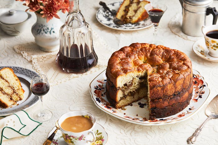 An opened aranygaluska cake sits on a platter, surrounded by cups of coffee, a bottle of cherry liqueur and a plate with a slice of cake.