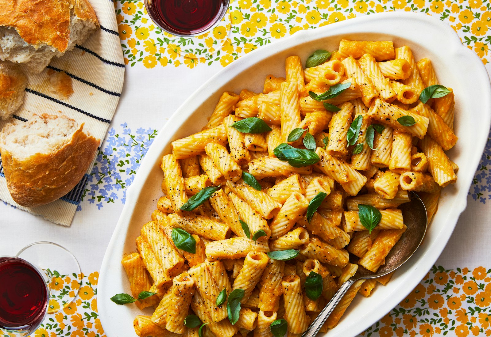 A bright orange butternut squash pasta with basil leaves in a serving dish, alongside cups of wine and Italian bread.