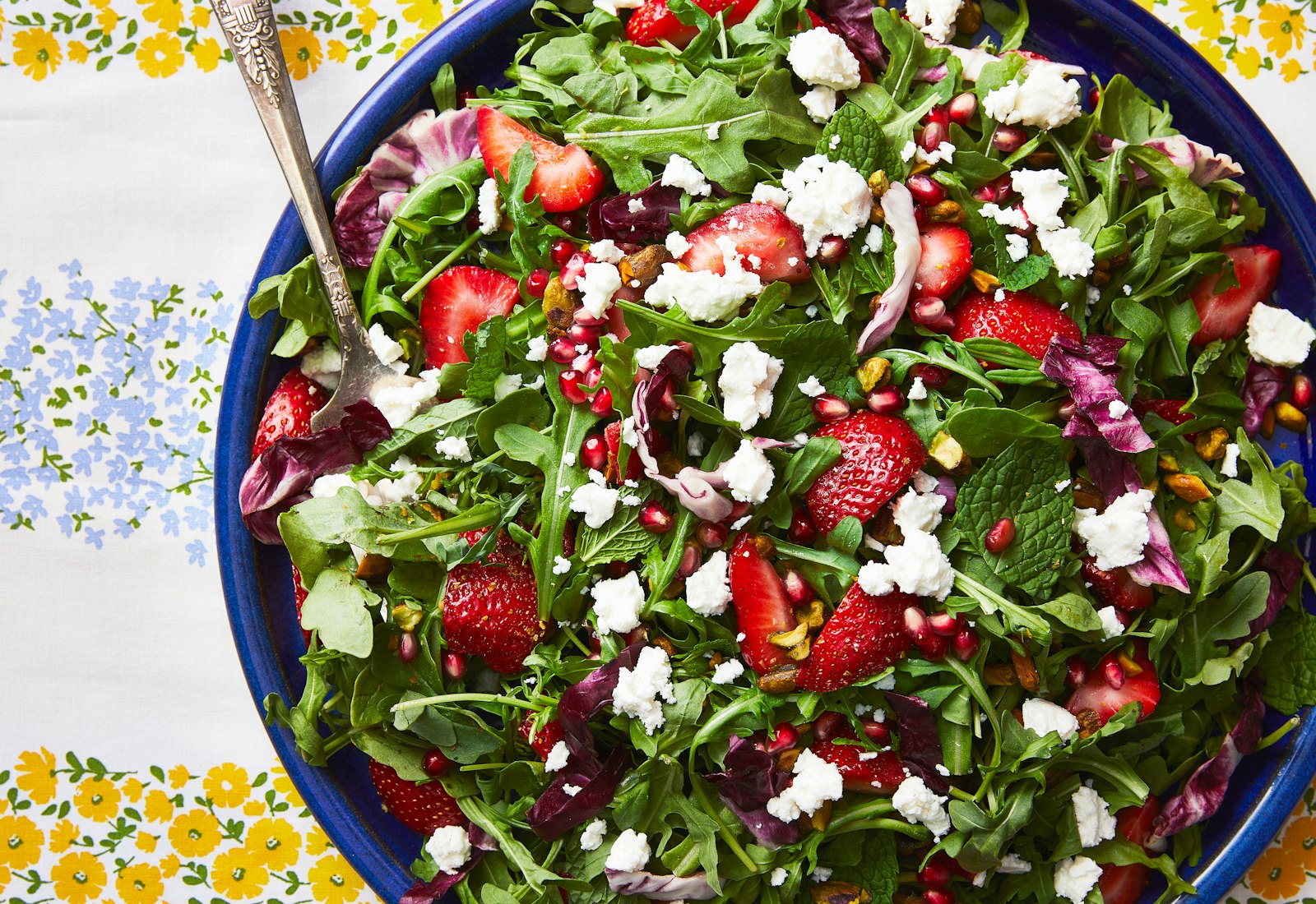 A colorful radicchio and arugula salad with strawberries, feta, pistachios and pomegranate on a striped tablecloth.