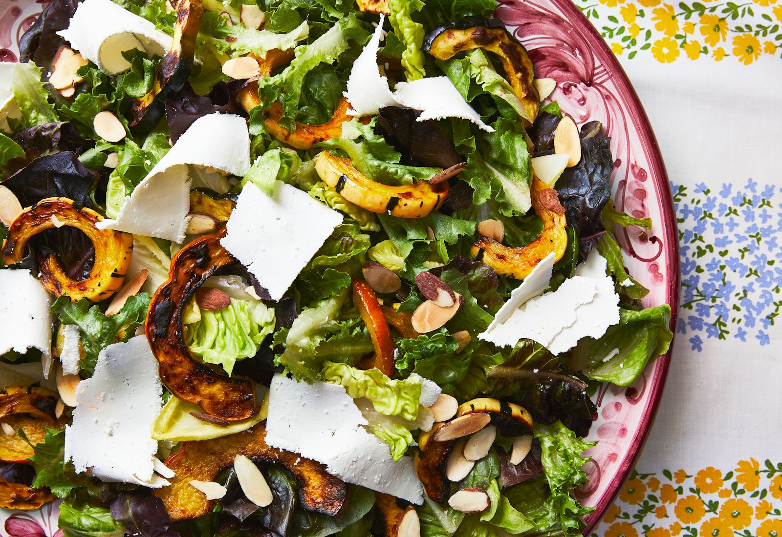 A bowl of colorful salad greens with roasted squash and ricotta salata on a printed tablecloth. 