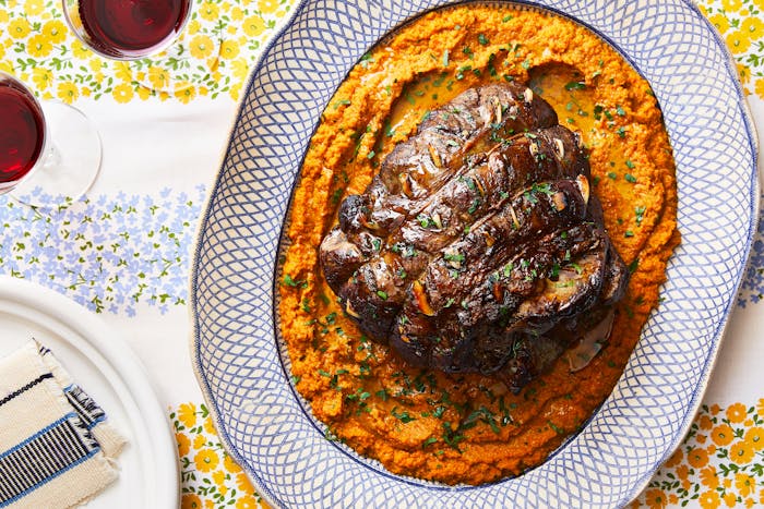 A roasted leg of lamb atop a vibrant orange vegetable spread, set on a platter. 