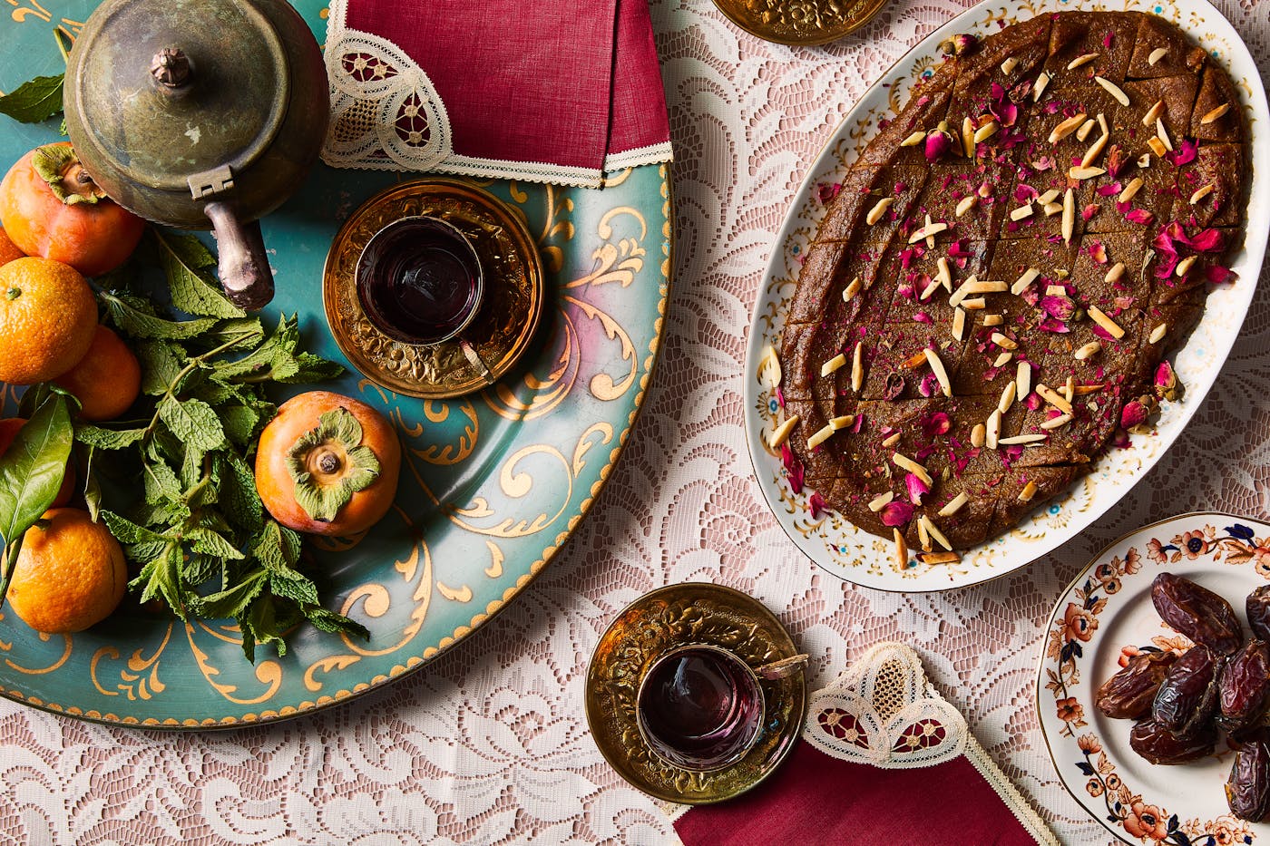 An oval platter of golden Persian halva covered with slivered almonds and crushed rose, surrounded by persimmon fruit and glasses of tea.