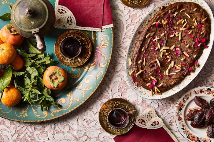 An oval platter of golden Persian halva covered with slivered almonds and crushed rose, surrounded by persimmon fruit and glasses of tea.