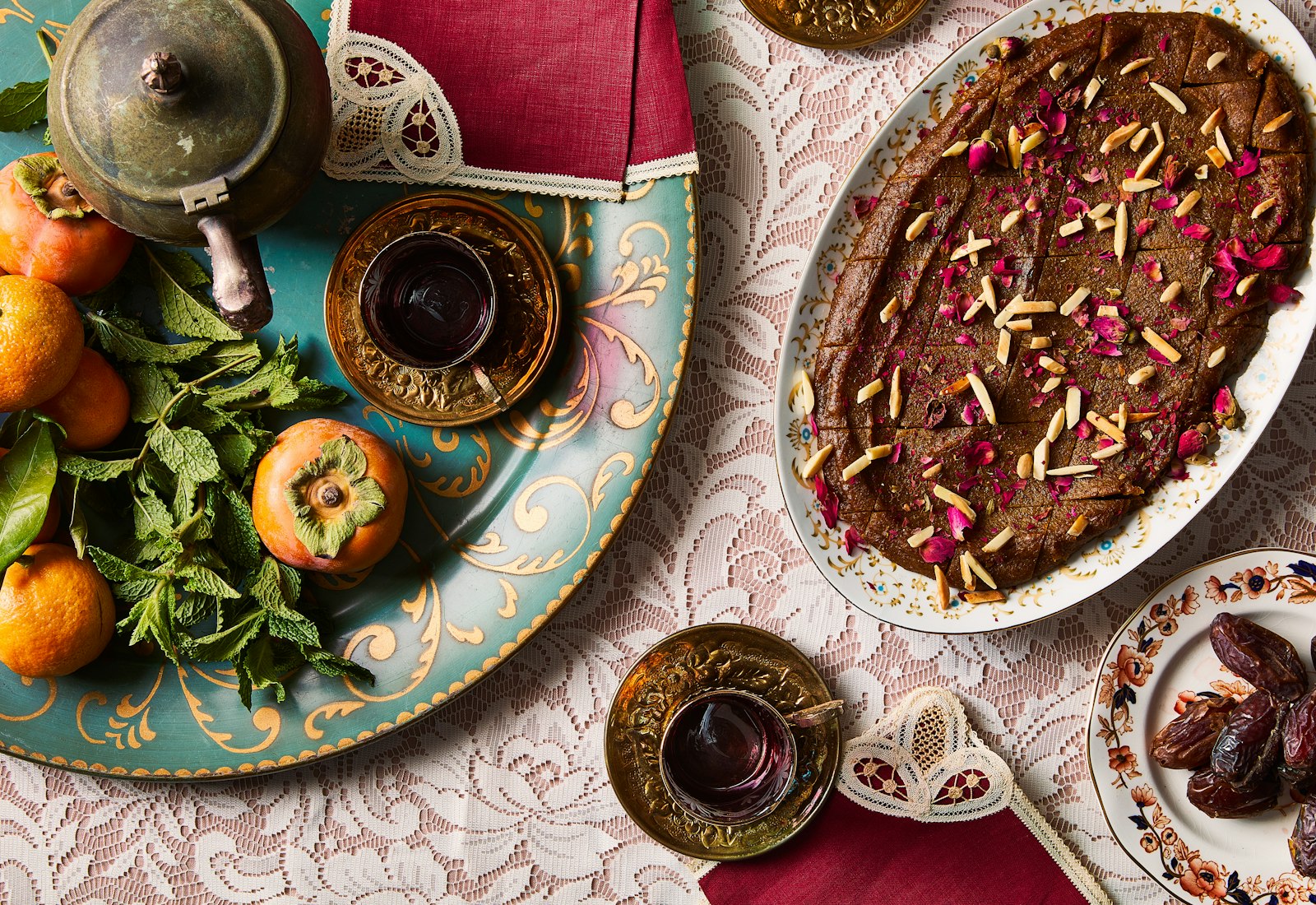 An oval platter of golden Persian halva covered with slivered almonds and crushed rose, surrounded by persimmon fruit and glasses of tea.