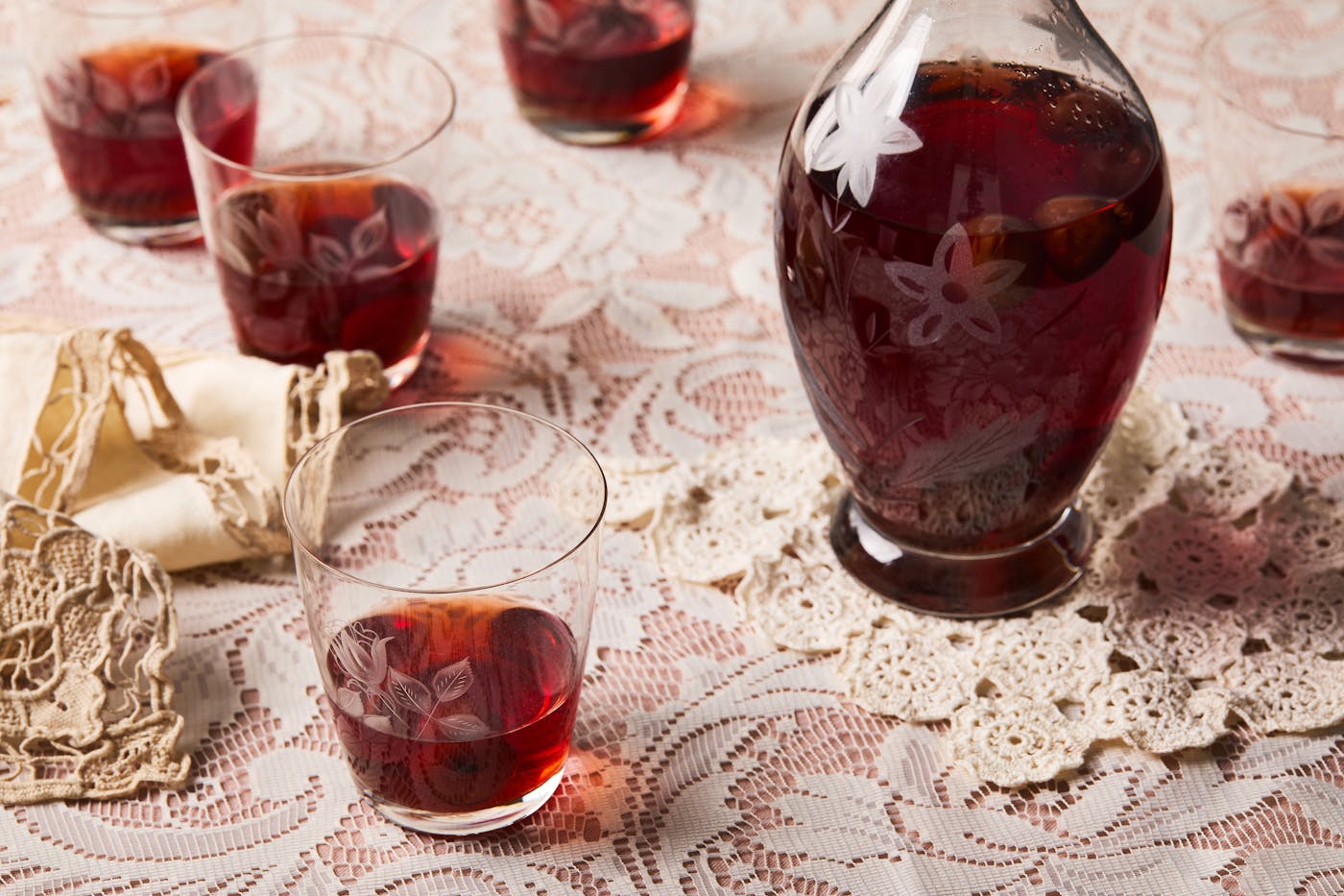 Shot glasses of bright red sour cherry infused vodka on a lacy, light pink tablecloth.