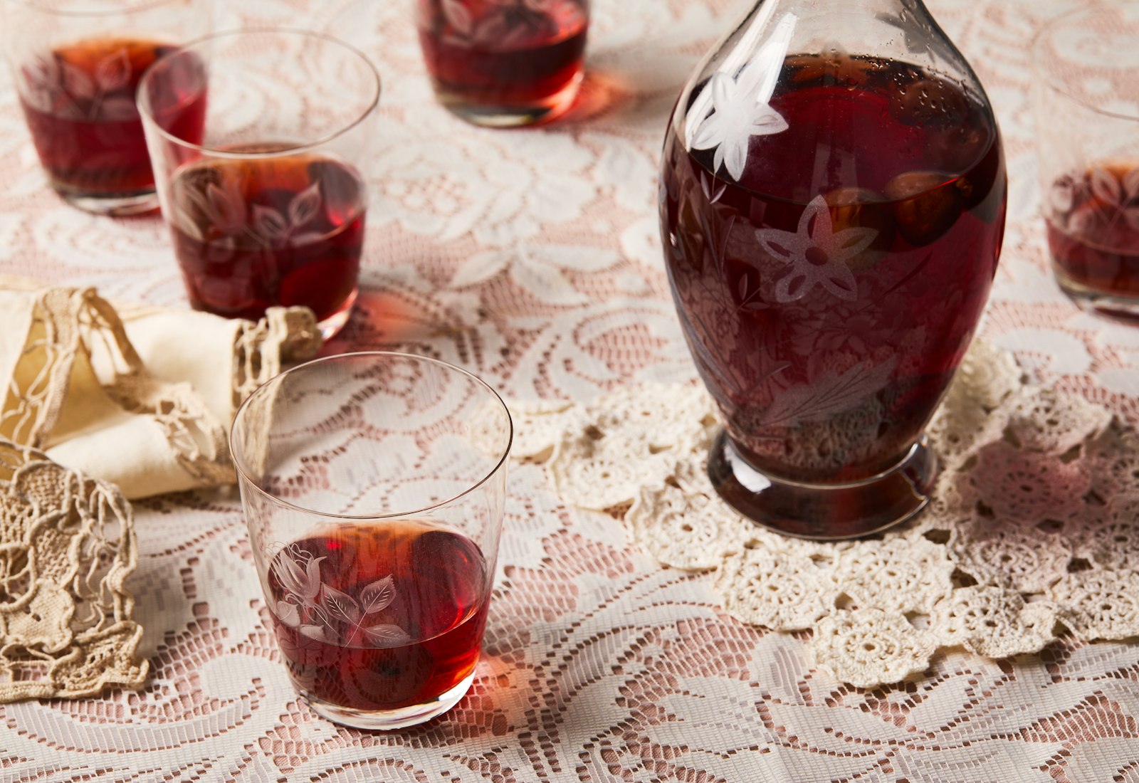 Shot glasses of bright red sour cherry infused vodka on a lacy, light pink tablecloth.