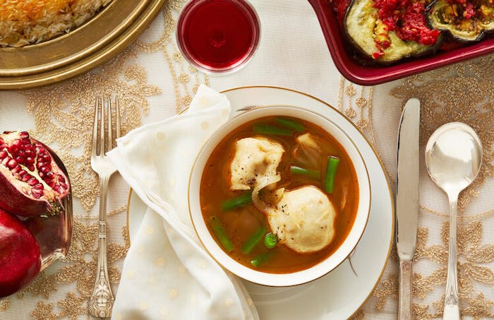 Dumpling soup with fresh cracked pepper, halved pomegranates, red wine atop formal tablecloth. 