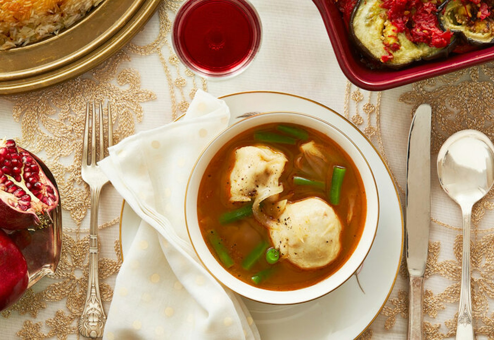 Dumpling soup with fresh cracked pepper, halved pomegranates, red wine atop formal tablecloth.