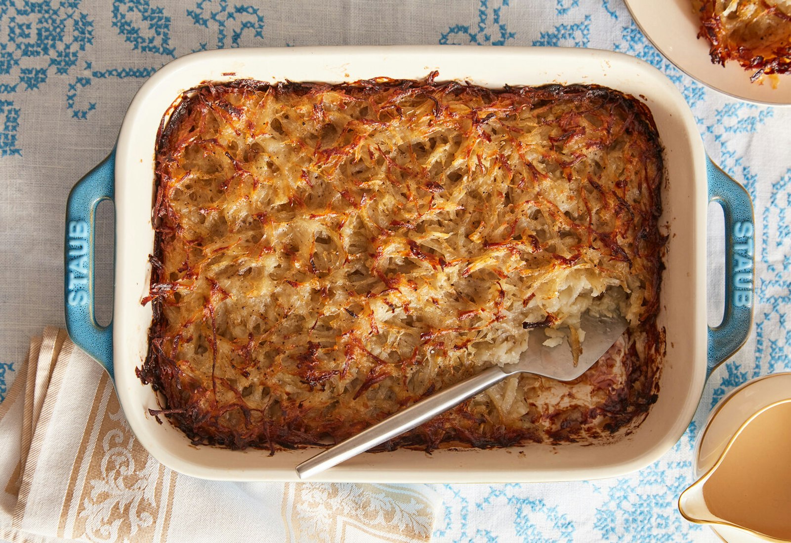 Kugel in blue and white casserole dish alongside pitcher of gravy atop blue embroidered tablecloth.