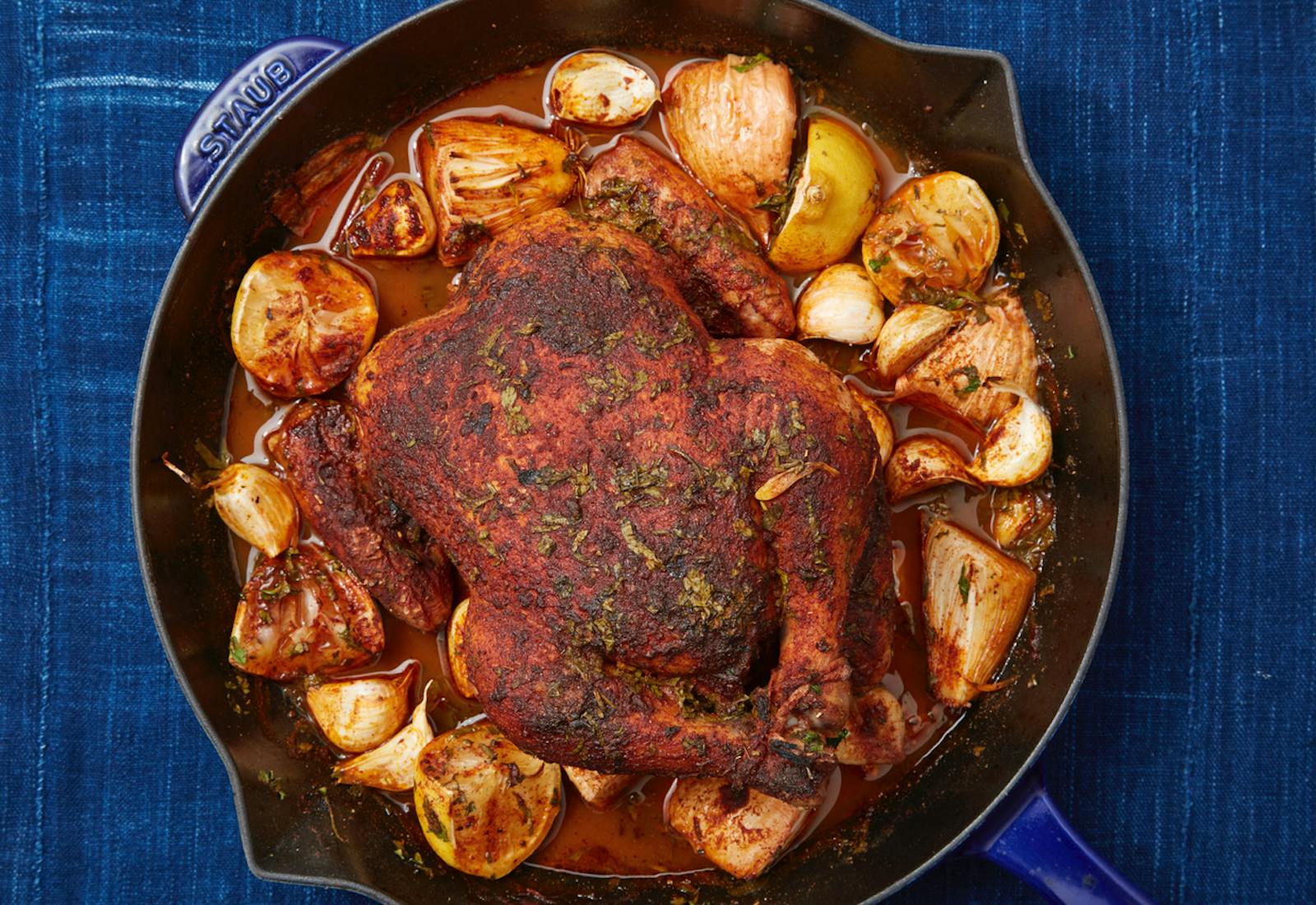 Spiced roasted chicken surrounded by lemon, onions and garlic in cast iron skillet atop denim tablecloth.