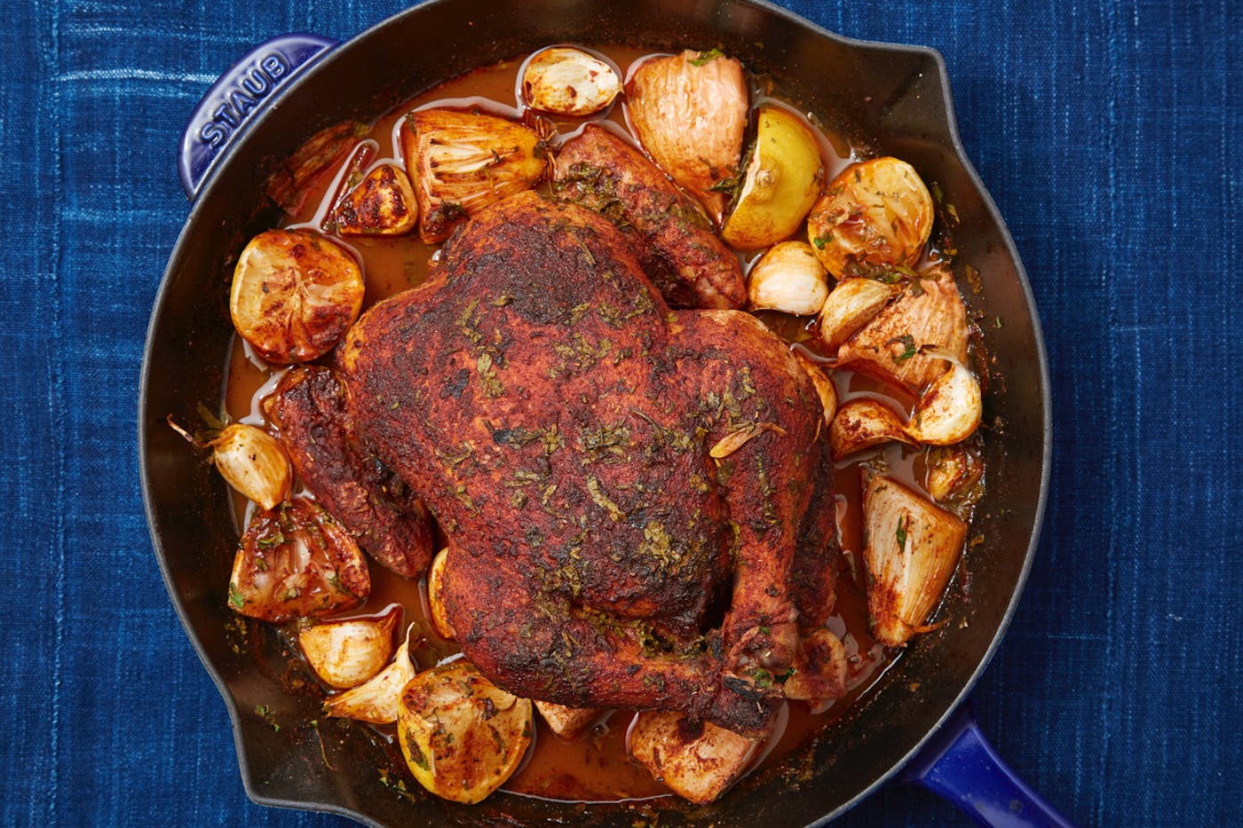 Spiced roasted chicken surrounded by lemon, onions and garlic in cast iron skillet atop denim tablecloth.