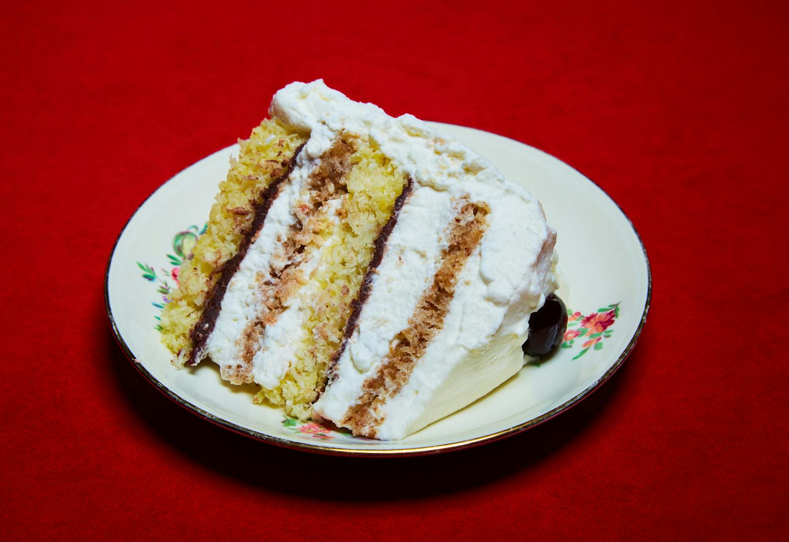 Slice of coconut macaroon layer cake on small floral dish atop red tablecloth.