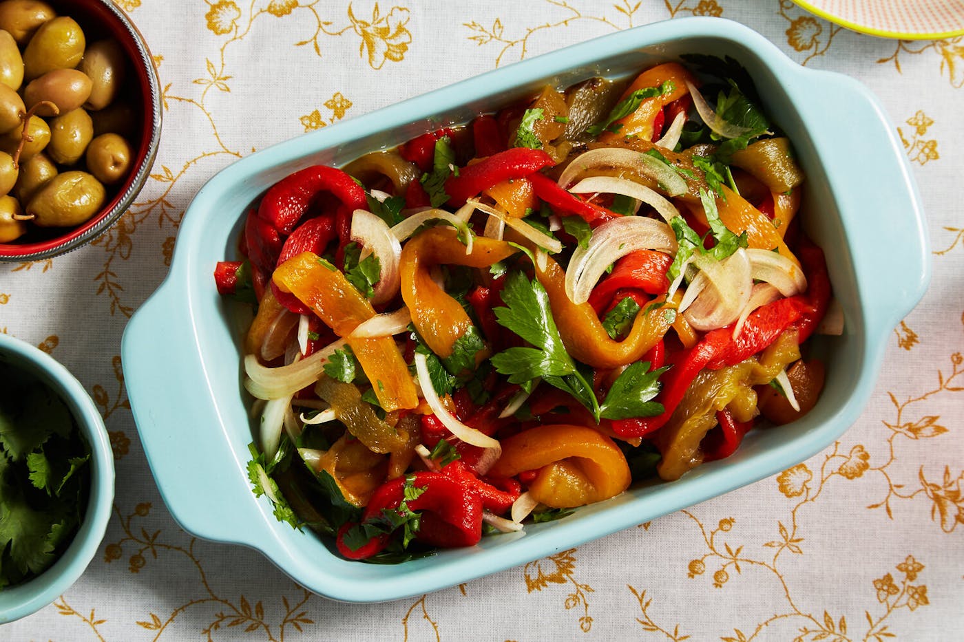 Colorful roasted pepper salad with parsley in blue casserole dish alongside bowl of olives, atop yellow floral tablecloth.