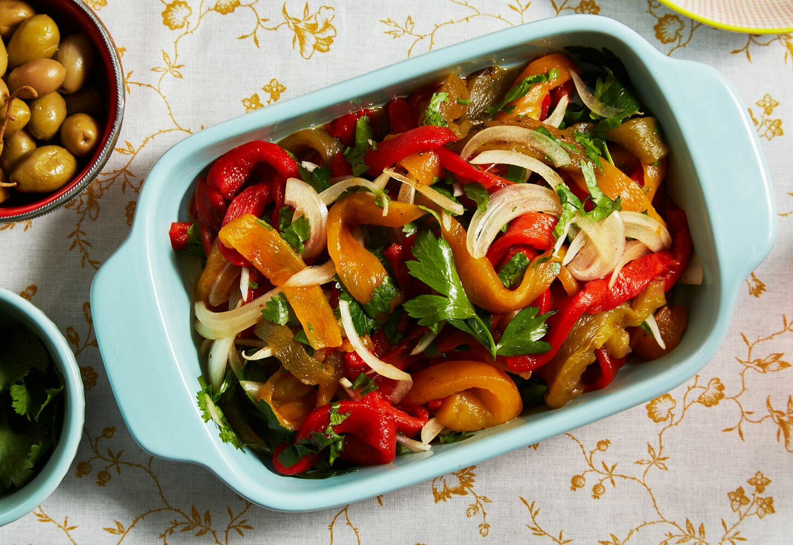 Colorful roasted pepper salad with parsley in blue casserole dish alongside bowl of olives, atop yellow floral tablecloth.