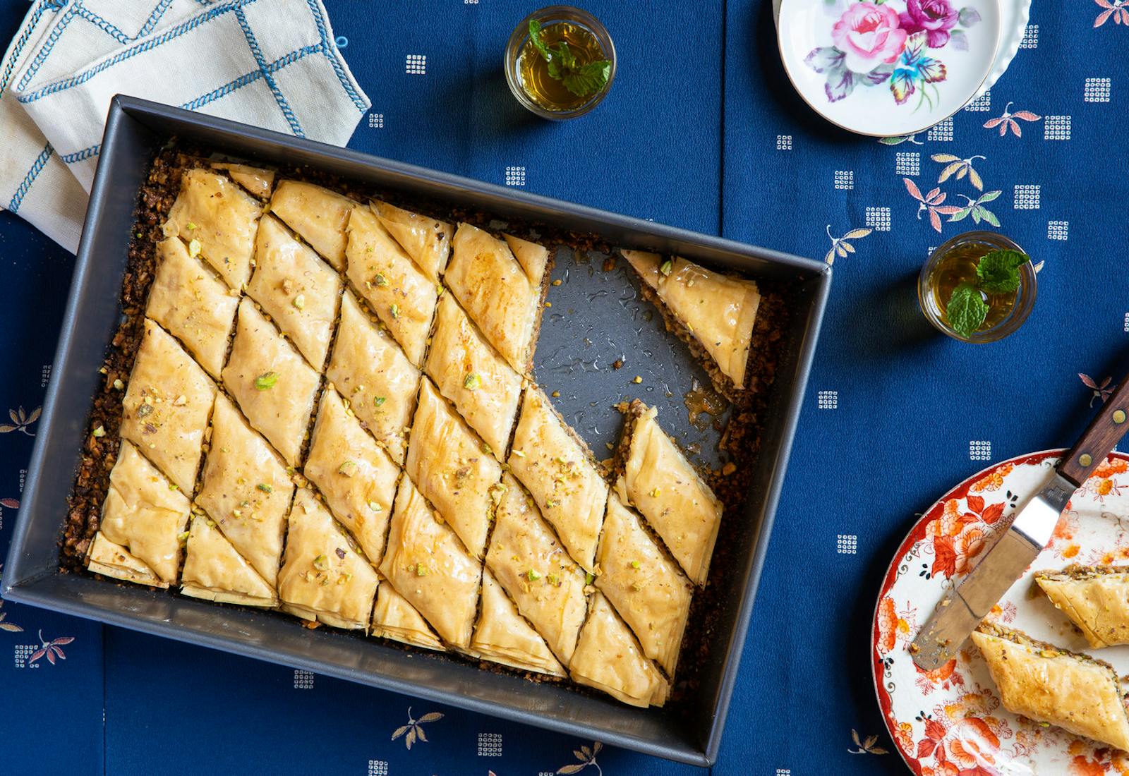 Sliced baklava in pan alongside mint tea, printed napkin and floral serving dishes, atop blue tablecloth.