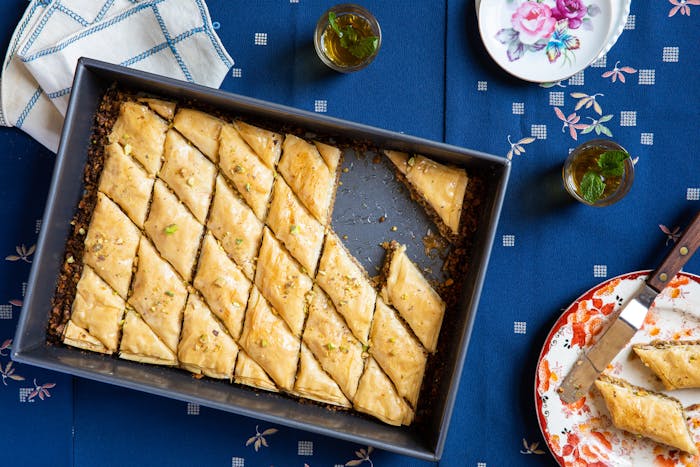 Sliced baklava in pan alongside mint tea, printed napkin and floral serving dishes, atop blue tablecloth.