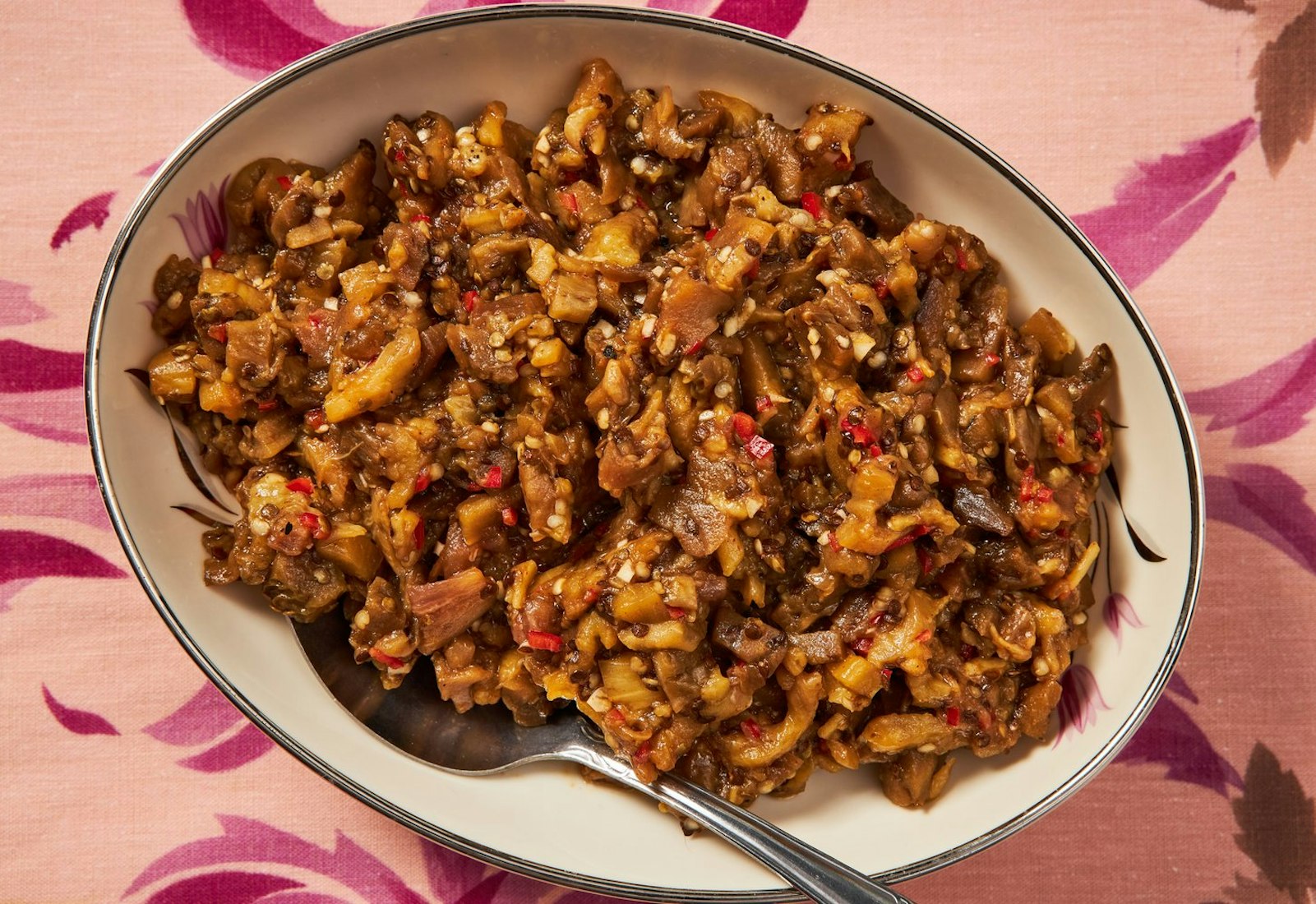 Roasted eggplant salad in large bowl atop pink floral tablecloth.
