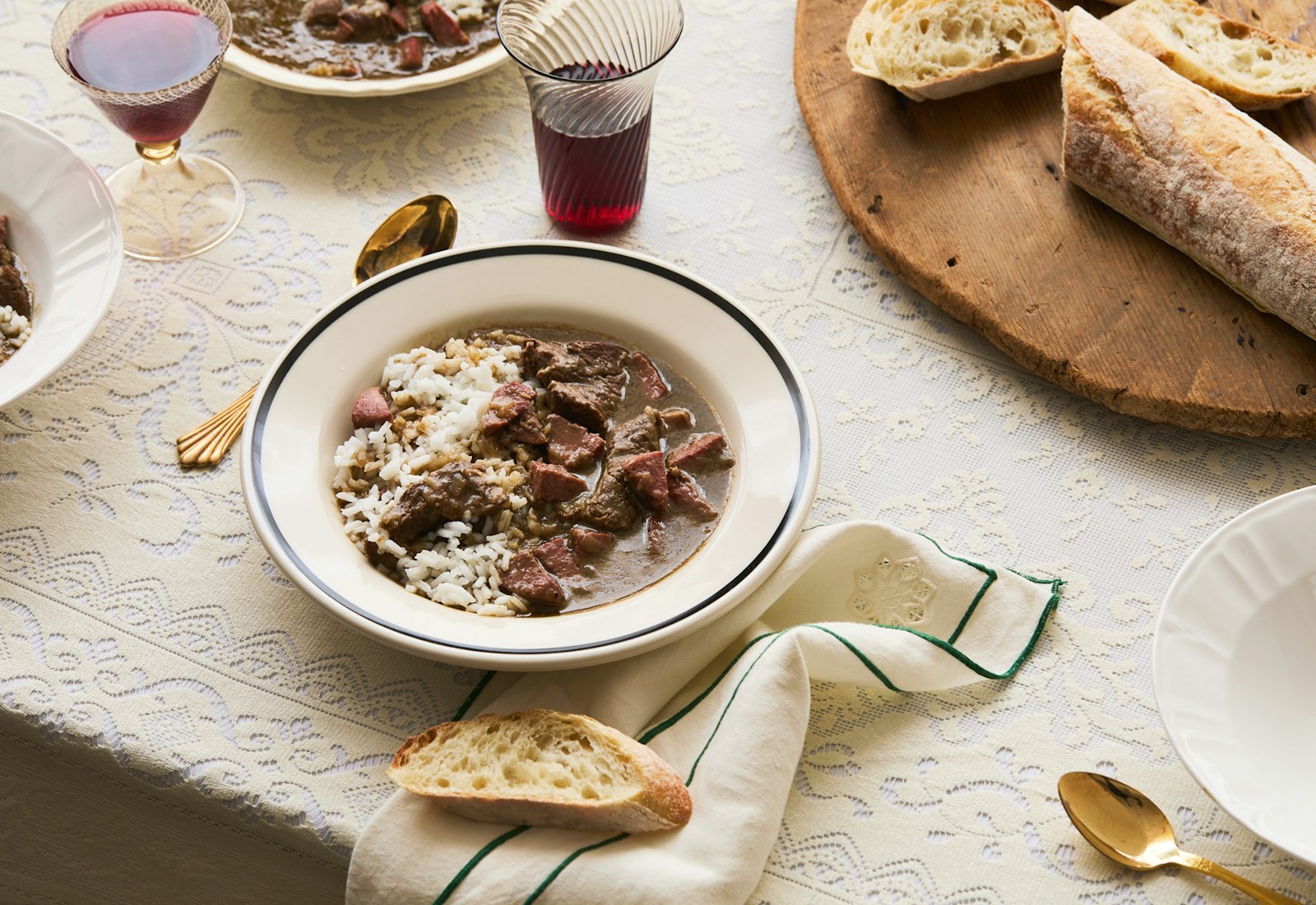 gumbo in a white bowl, french bread on cutting board and glasses of red wine