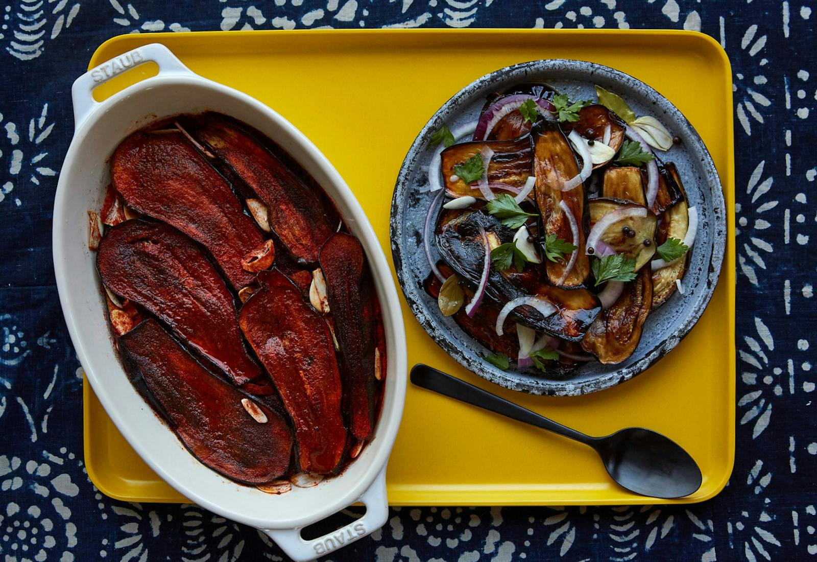 Eggplant in paprika in white casserole dish next to eggplant in vinegar on blue plate on yellow tray, atop blue patterned tablecloth.