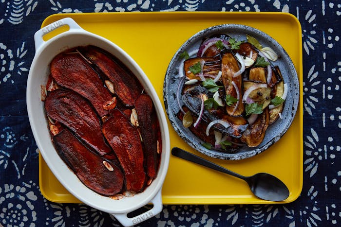 Eggplant in paprika in white casserole dish next to eggplant in vinegar on blue plate on yellow tray, atop blue patterned tablecloth.