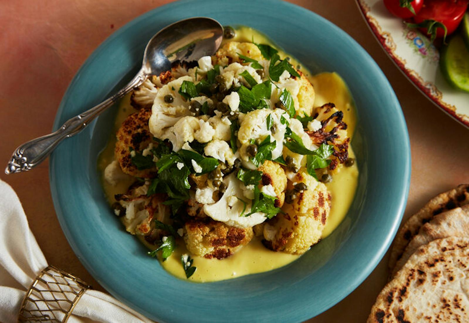 Roasted cauliflower over dijon sauce with fresh parsley alongside pita and plate of sliced cucumbers and tomatoes.
