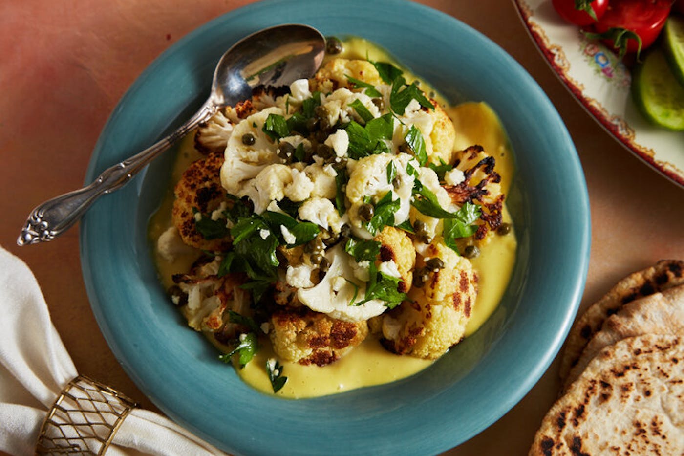 Roasted cauliflower over dijon sauce with fresh parsley alongside pita and plate of sliced cucumbers and tomatoes.