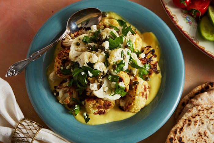 Roasted cauliflower over dijon sauce with fresh parsley alongside pita and plate of sliced cucumbers and tomatoes.