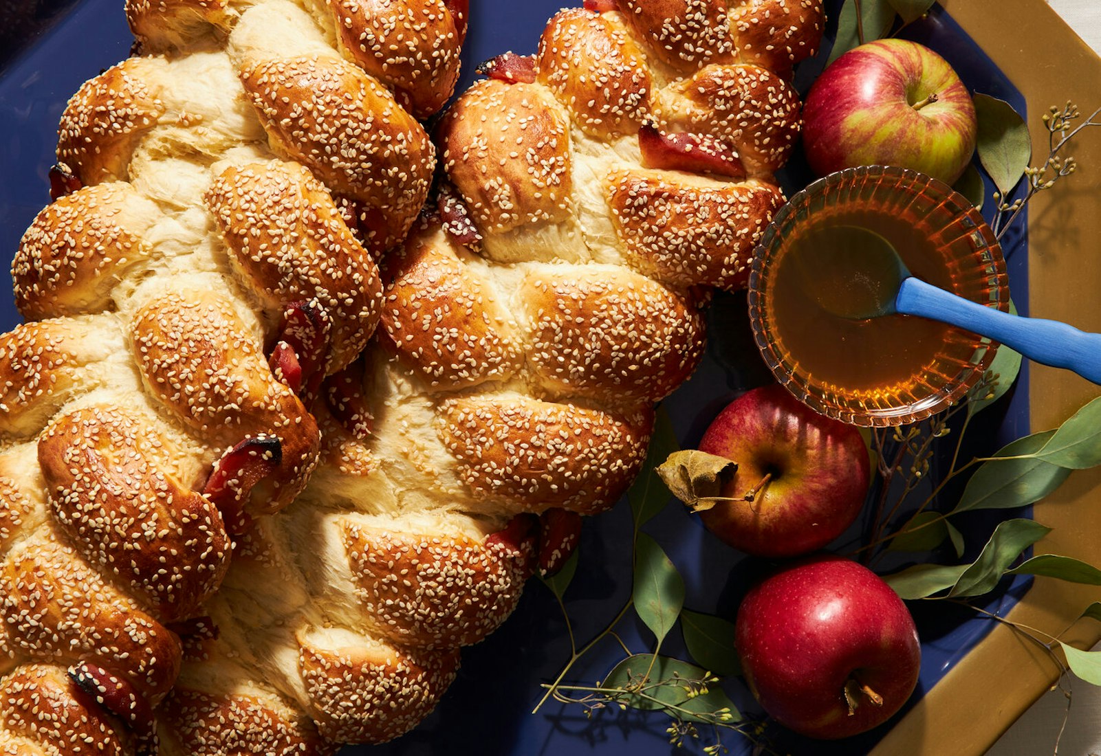 Sweet challah with bowl of honey and apples with leaves atop blue and gold platter.
