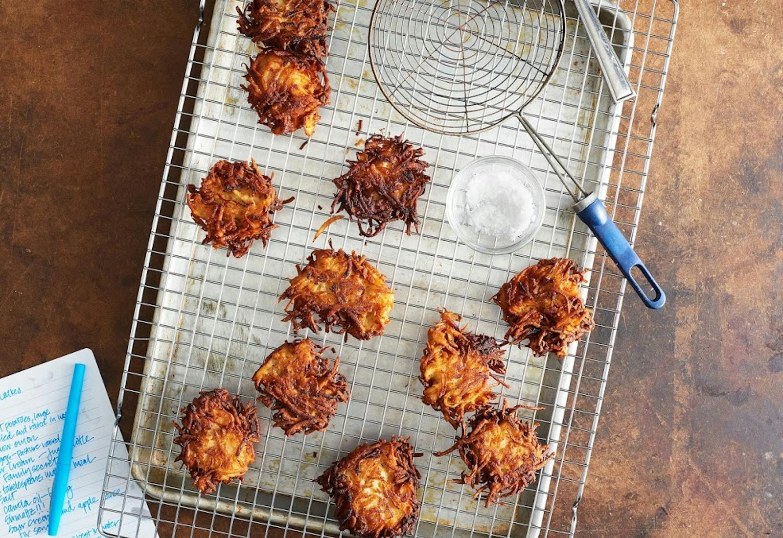 Fried latkes draining on metal rack with dish of coarse salt and handwritten recipe atop brown surface. 
