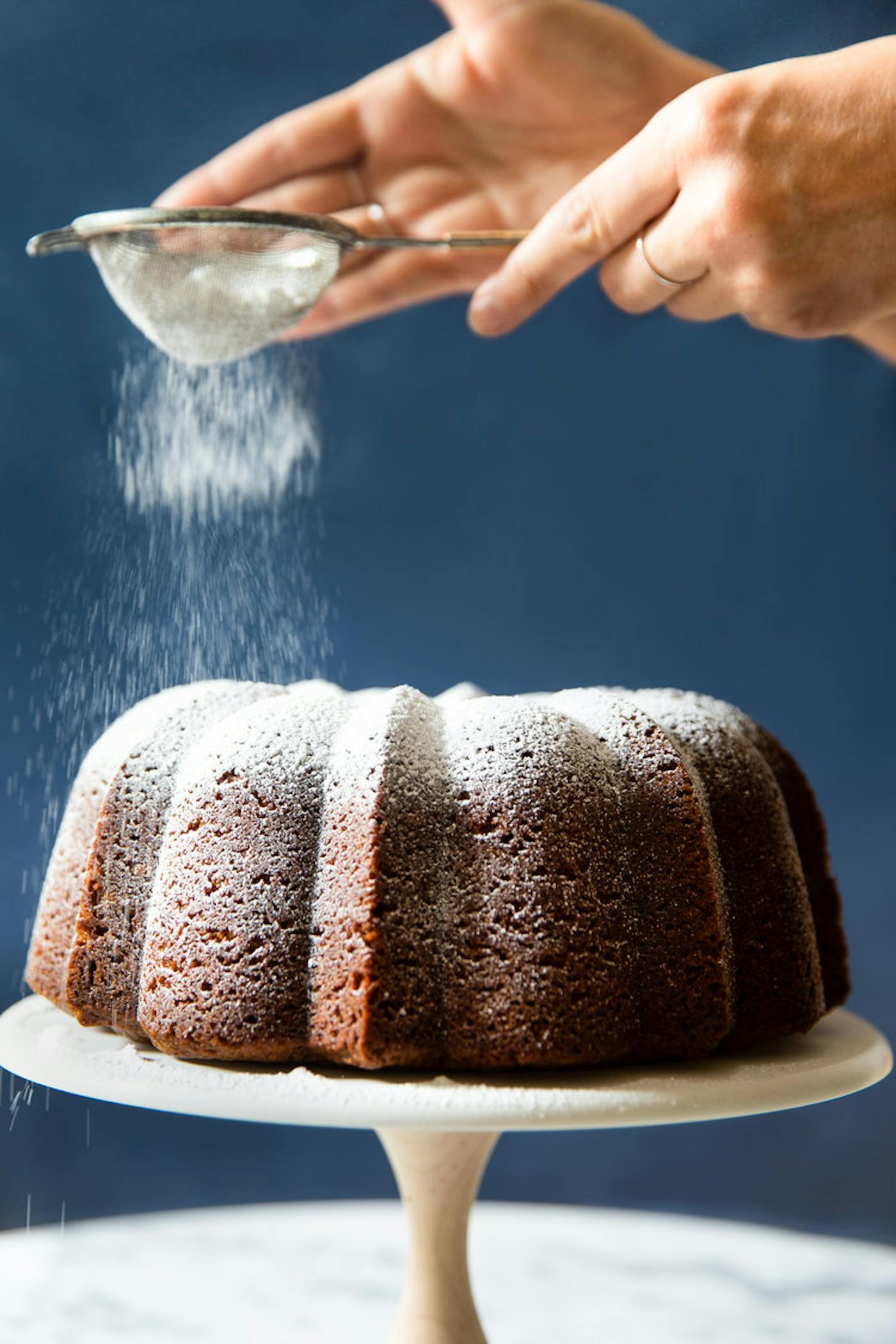 Chef dusting powdered sugar onto caramelized honey cake, on white cake stand.