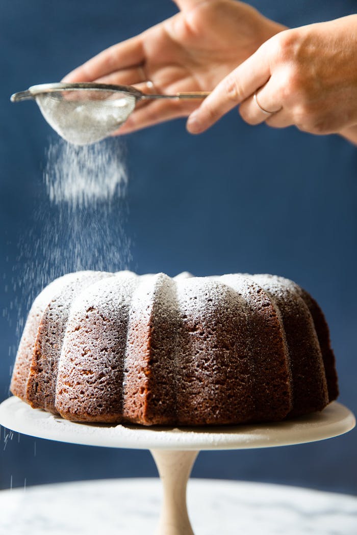 Chef dusting powdered sugar onto caramelized honey cake, on white cake stand.