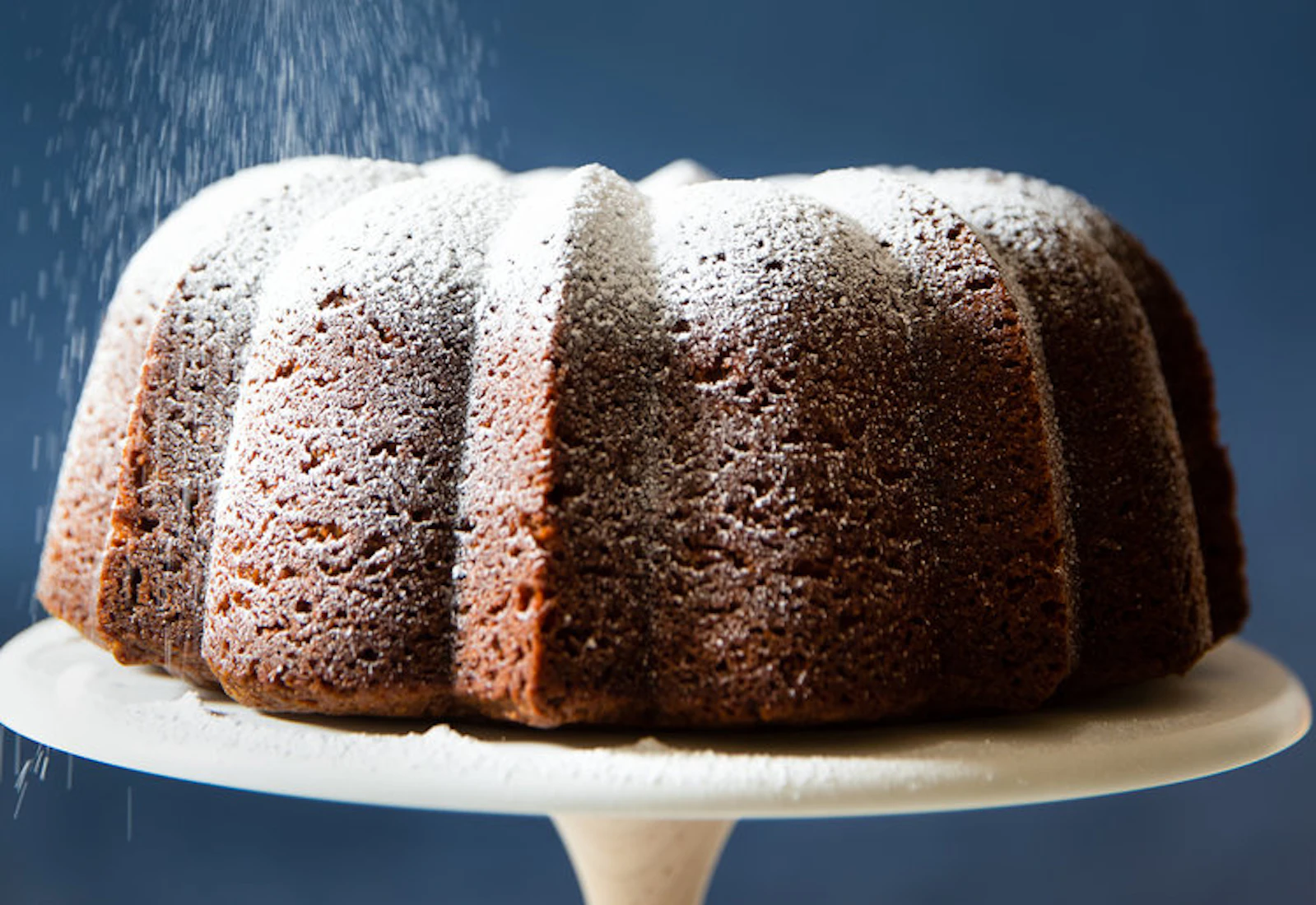 Chef dusting powdered sugar onto caramelized honey cake, on white cake stand.