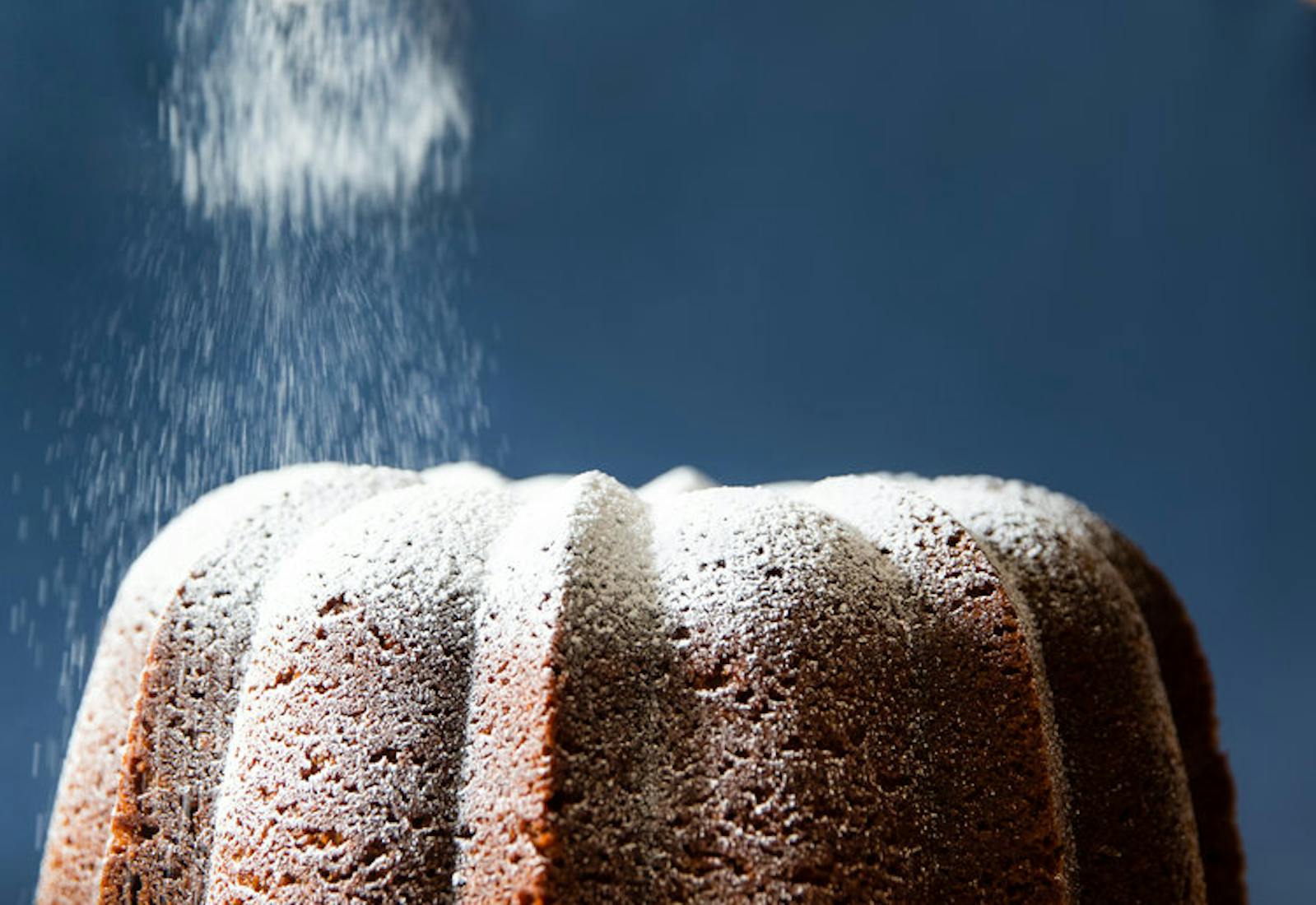 Chef dusting powdered sugar onto caramelized honey cake, on white cake stand.