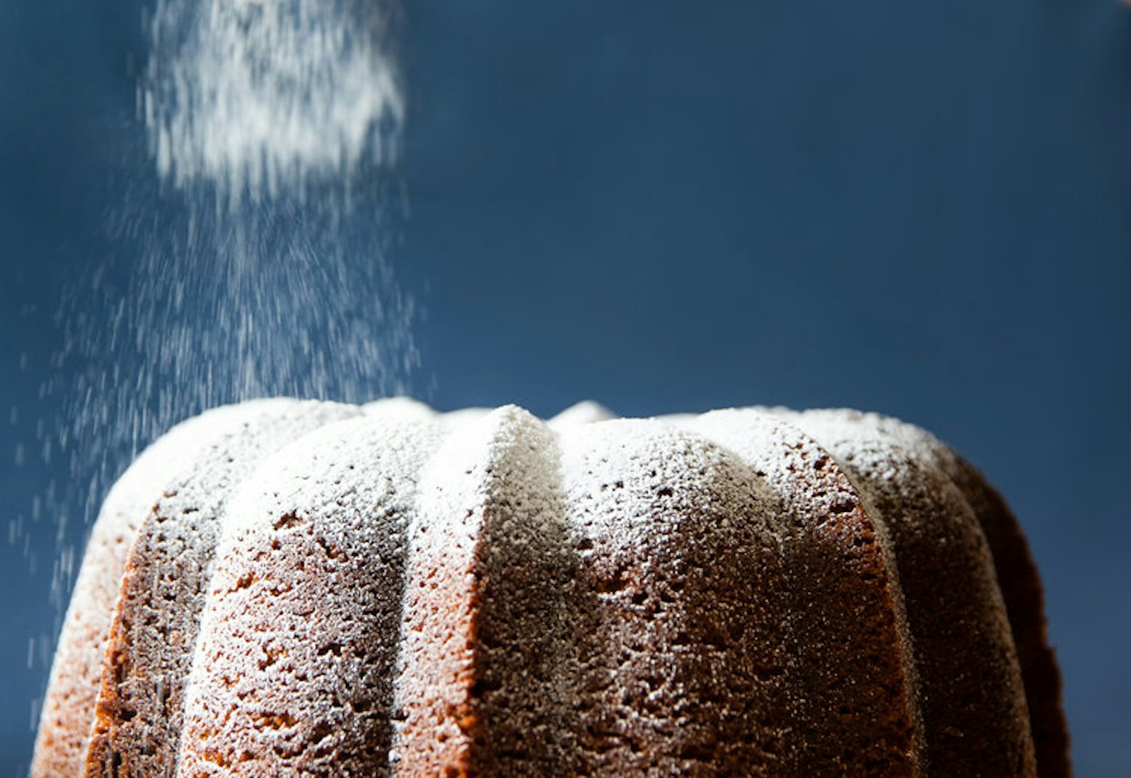 Chef dusting powdered sugar onto caramelized honey cake, on white cake stand.