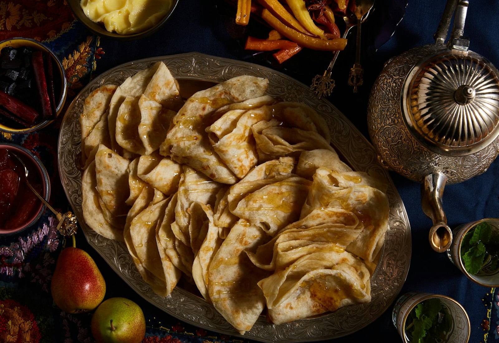 Mufleta layered onto large gold platter drizzled with honey alongside bowl of softened butter, a mint tea kettle, bowls of spices, and pears.