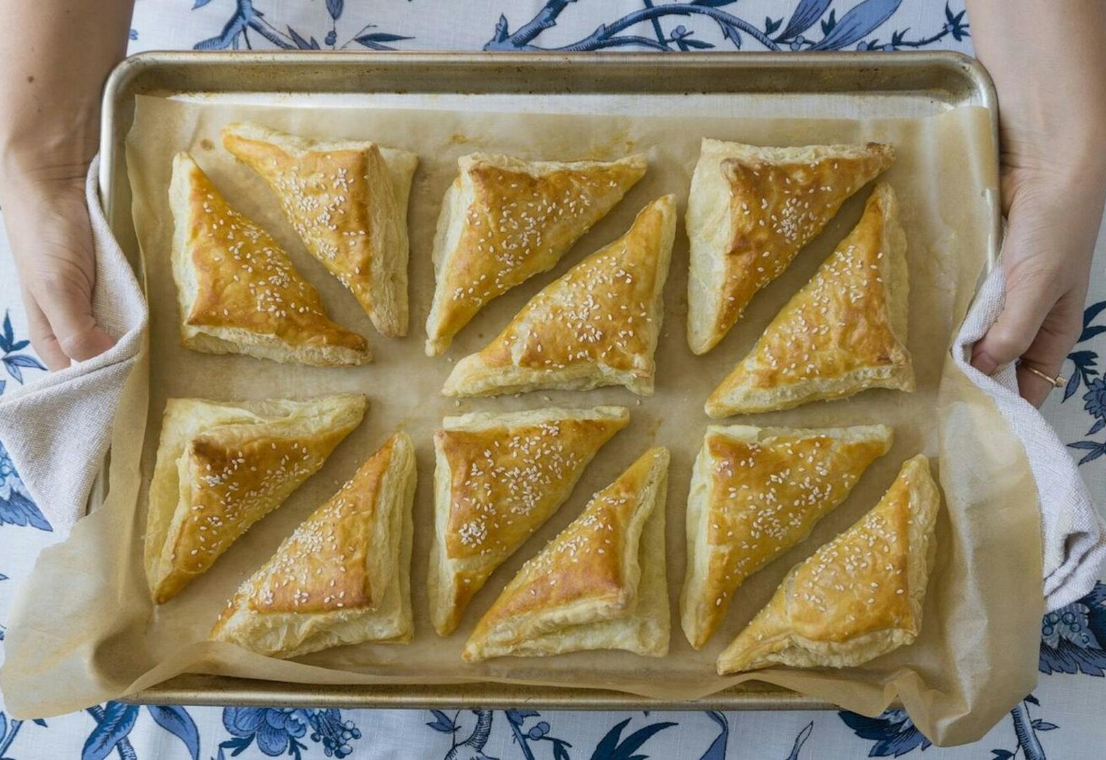 Chef holding tray of freshly baked Turkish bourekas atop blue printed tablecloth. 