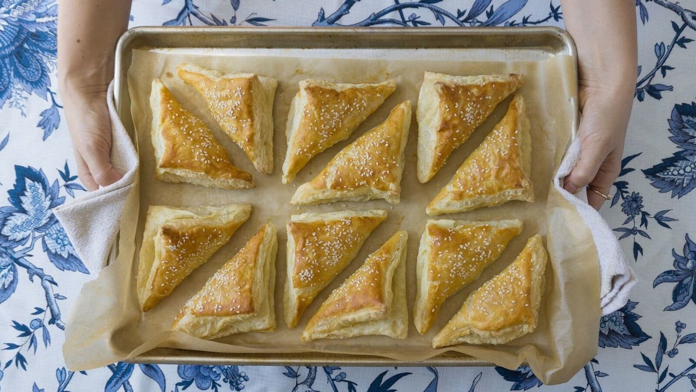Chef holding tray of freshly baked Turkish bourekas atop blue printed tablecloth. 