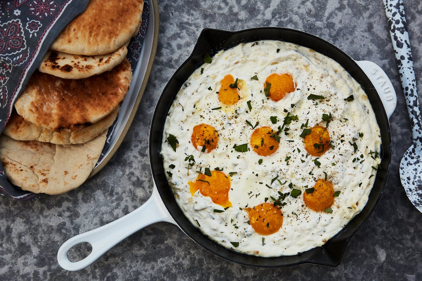 White shakshuka with labneh in cast iron skilled alongside fresh pita, atop speckled grey surface.