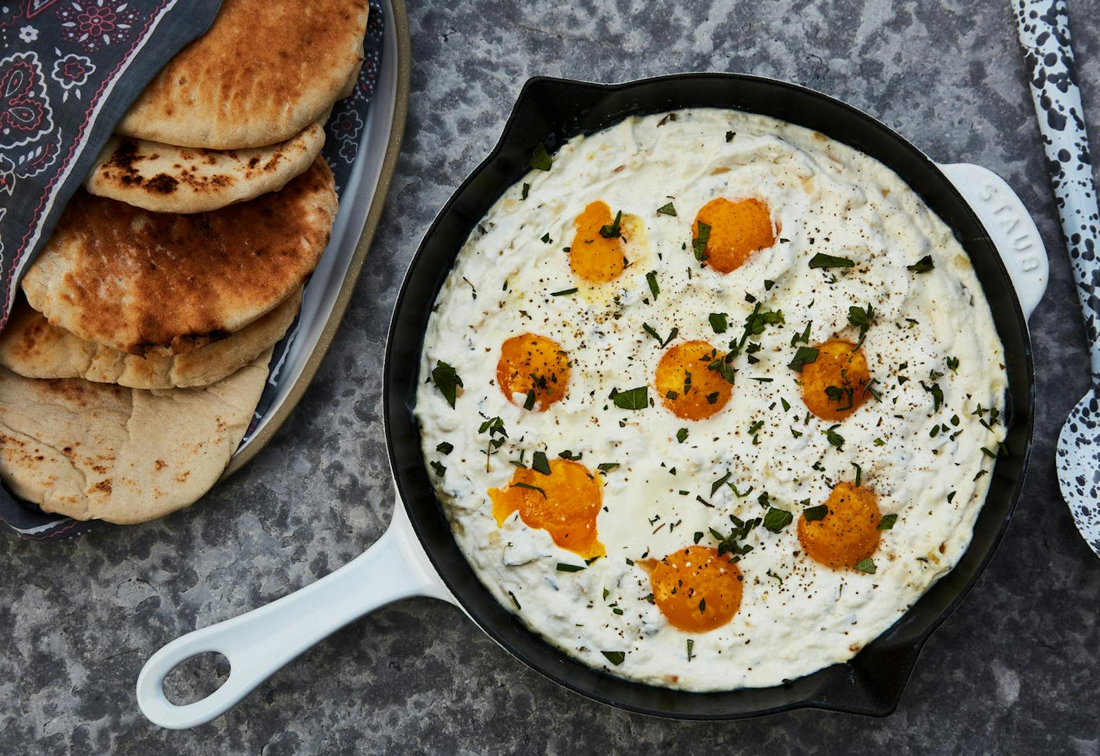 White shakshuka with labneh in cast iron skilled alongside fresh pita, atop speckled grey surface.