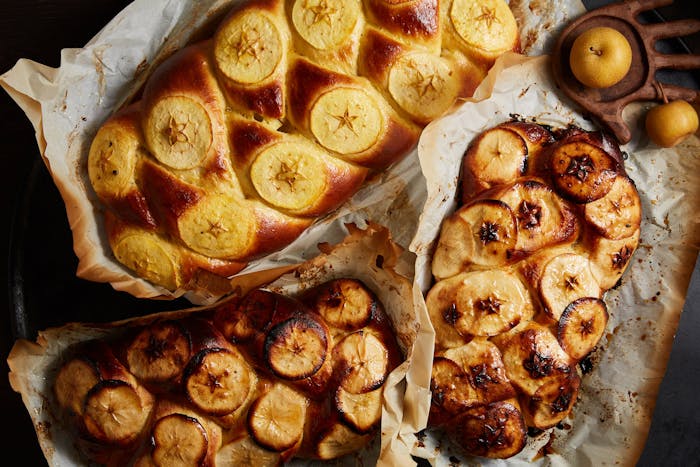 Loaves of challah woven with sliced apples on browned parchment paper alongside fresh apples.