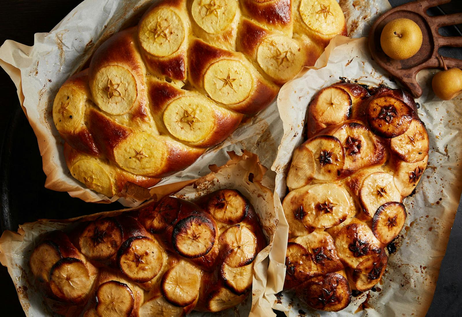Loaves of challah woven with sliced apples on browned parchment paper alongside fresh apples.