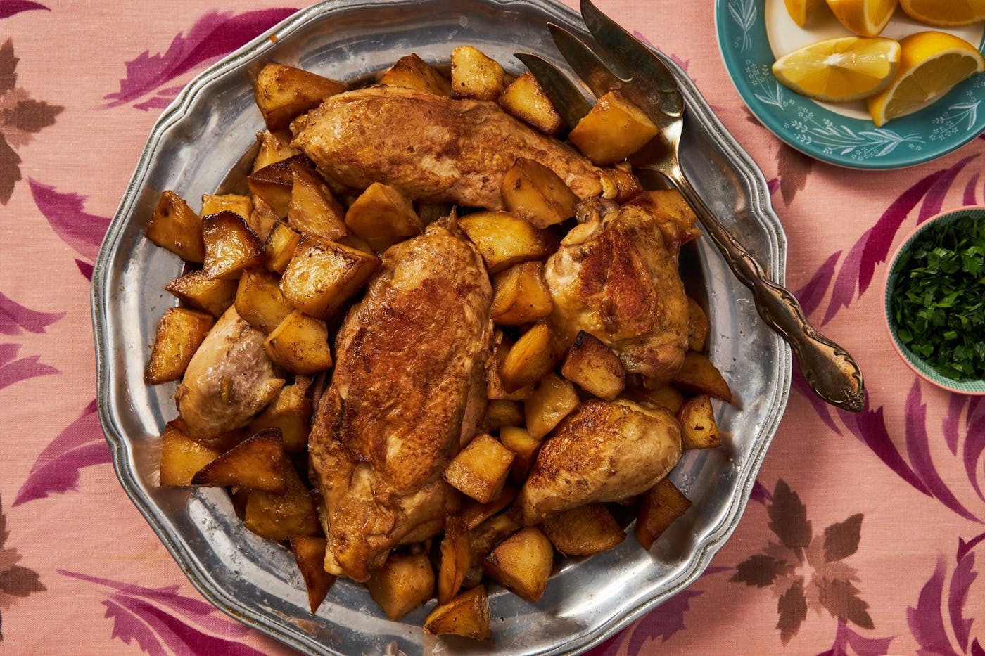 Chicken and potatoes on silver tray, lemons and parsley atop pink floral tablecloth.
