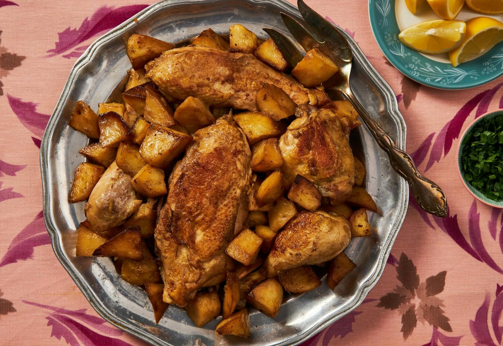 Chicken and potatoes on silver tray, lemons and parsley atop pink floral tablecloth.