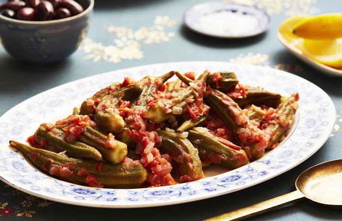 Okra with tomatoes, lemon wedges and olives atop navy tablecloth.